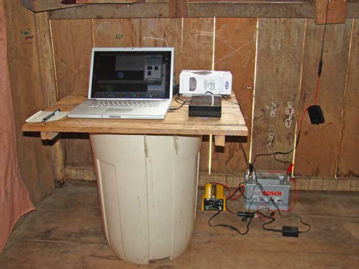A Mac laptop sits atop a wooden board that sits atop a plastic garbage can. The laptop receives power from a car battery through a yellow box. The car battery receives power from a cable that comes down from the roof. The laptop is connected to a hard drive.