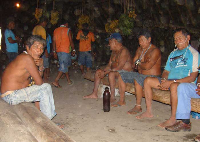 Elderly indigenous Amazonians sit on two parallel wooden benches inside a longhouse. One of the elders sings. In the background, young Marubo walk around the center of the longhouse. Bundles of bananas and plantains hang from the rafters.
